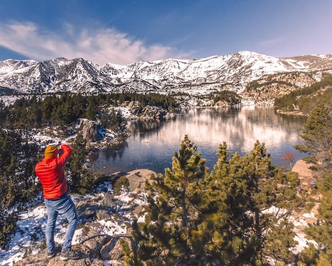 Un randonneur en veste rouge photographie un lac reflétant les sommets neigeux.